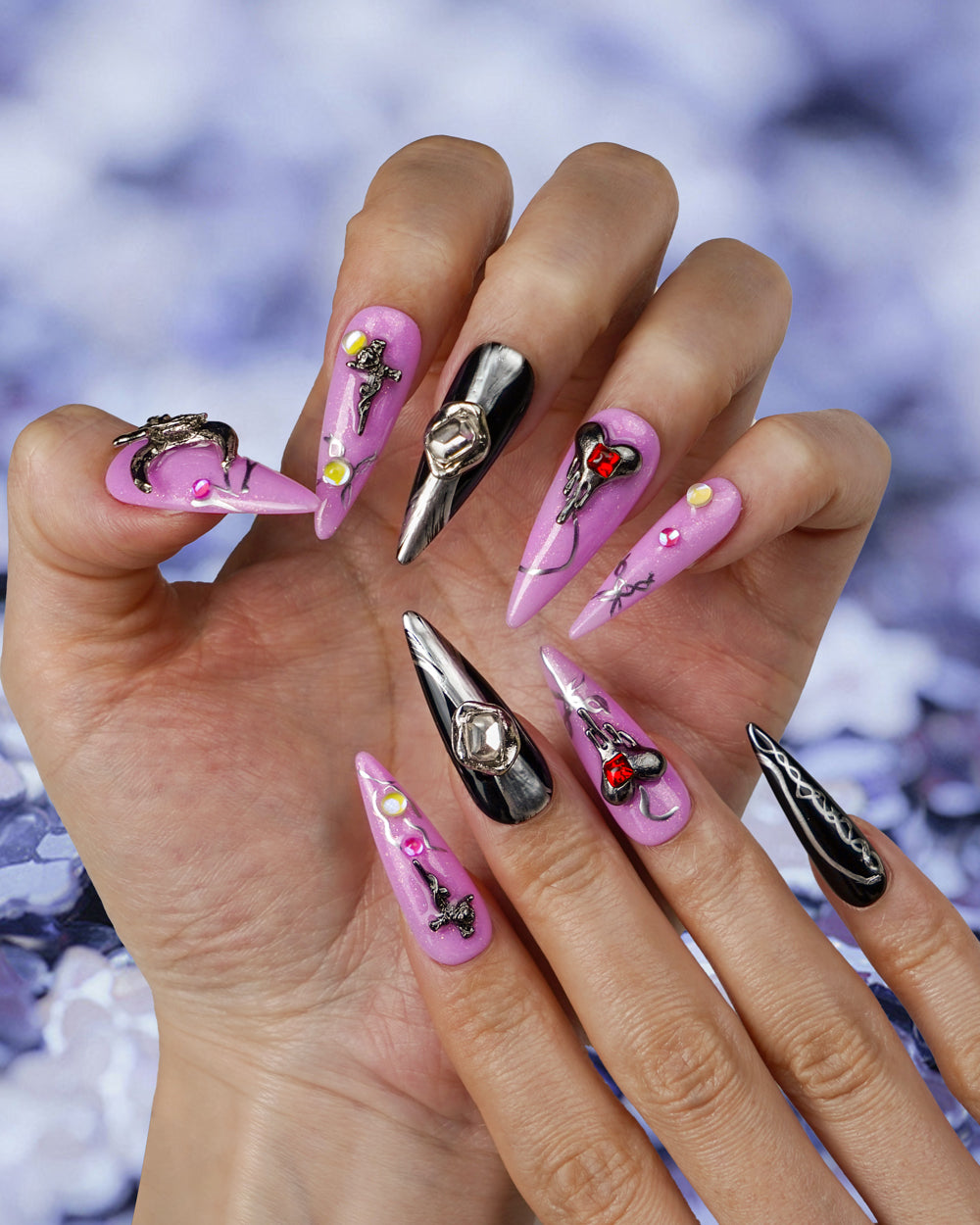 Trendy pink press-on nails with metallic accents, featuring intricate designs including crosses, heart-shaped gemstones, and black glossy nails with silver details.