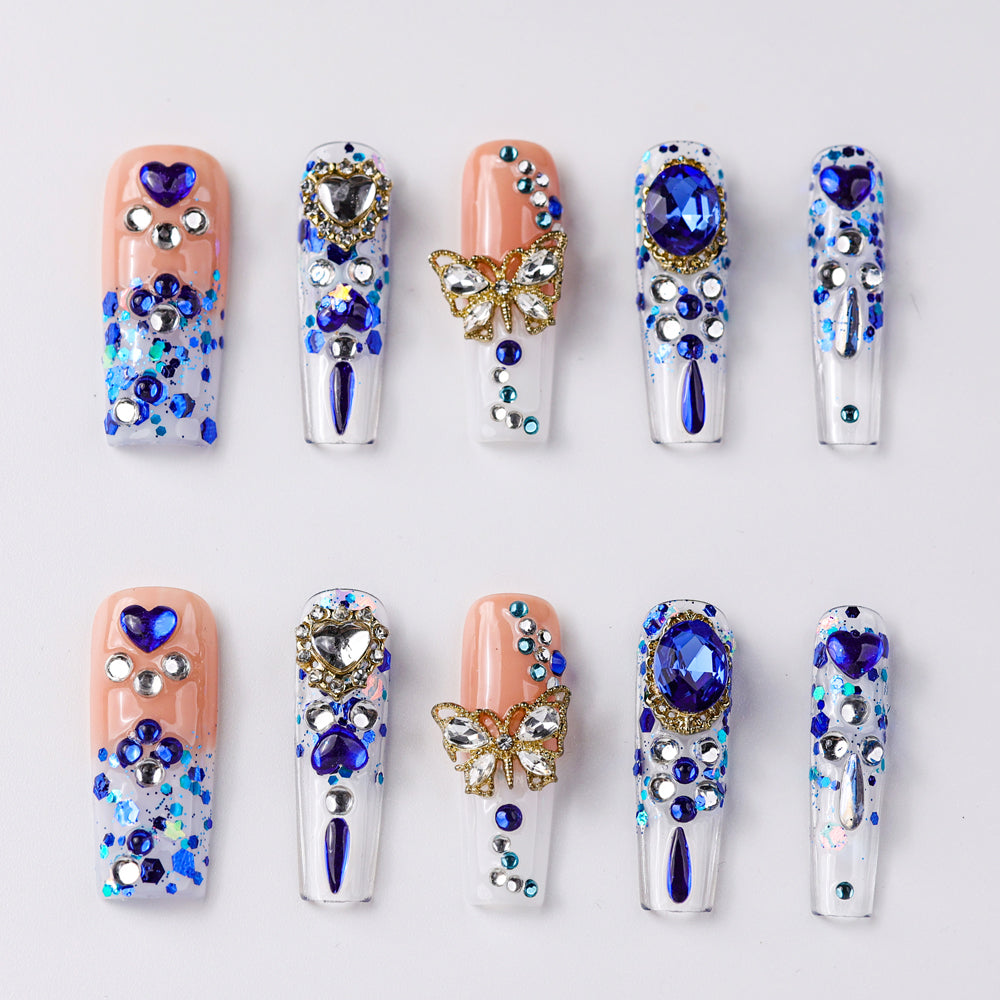 Vibrant press-on nails featuring a mix of blue and silver rhinestones, heart and butterfly accents, and scattered glitter designs. Bold, glamorous look with a combination of clear and soft peach nails.