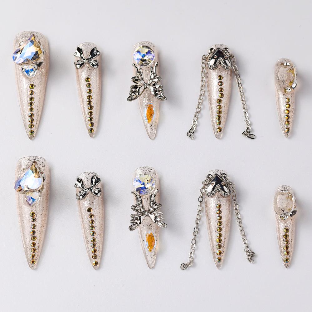 Elegant beige press-on nails adorned with rhinestones, bows, and metallic chain accents. Features intricate crystal designs and delicate embellishments for a chic and glamorous look.