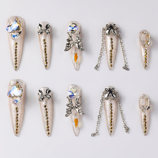 Elegant beige press-on nails adorned with rhinestones, bows, and metallic chain accents. Features intricate crystal designs and delicate embellishments for a chic and glamorous look.